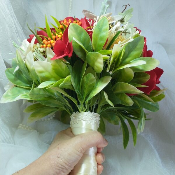 Buquê de flores com folhagem verde e detalhes vermelhos, mãos segurando o arranjo.