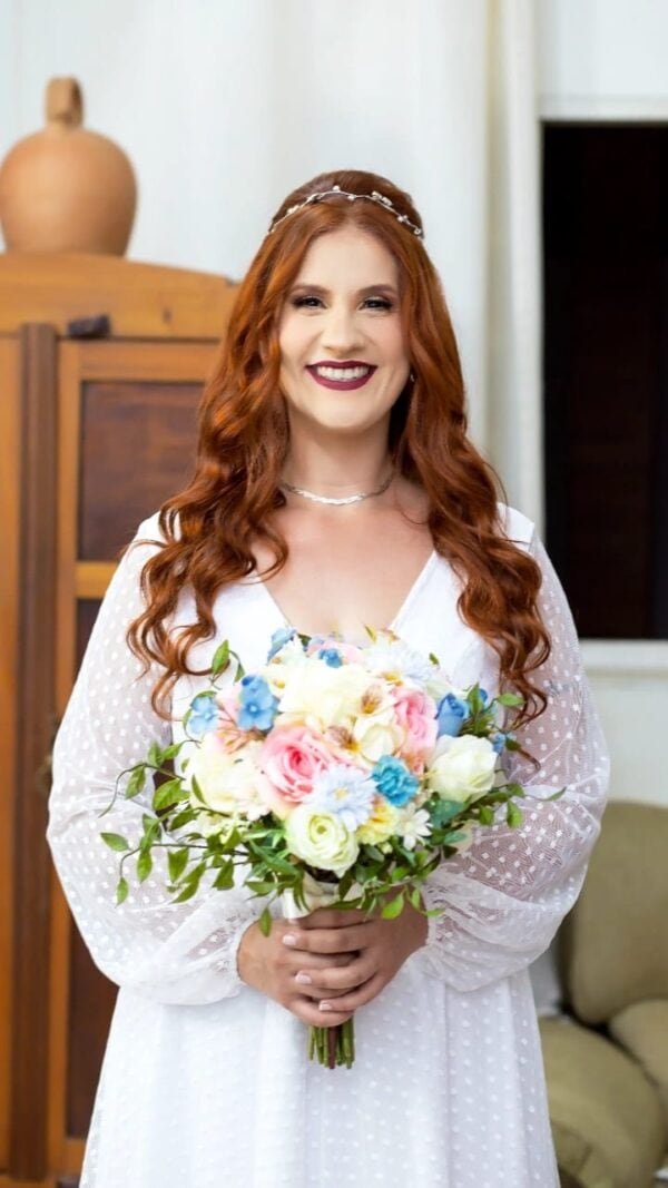 Bouquet de casamento com flores coloridas e folhagem, mulher sorridente, vestido branco, decoração de imóvel ao fundo.