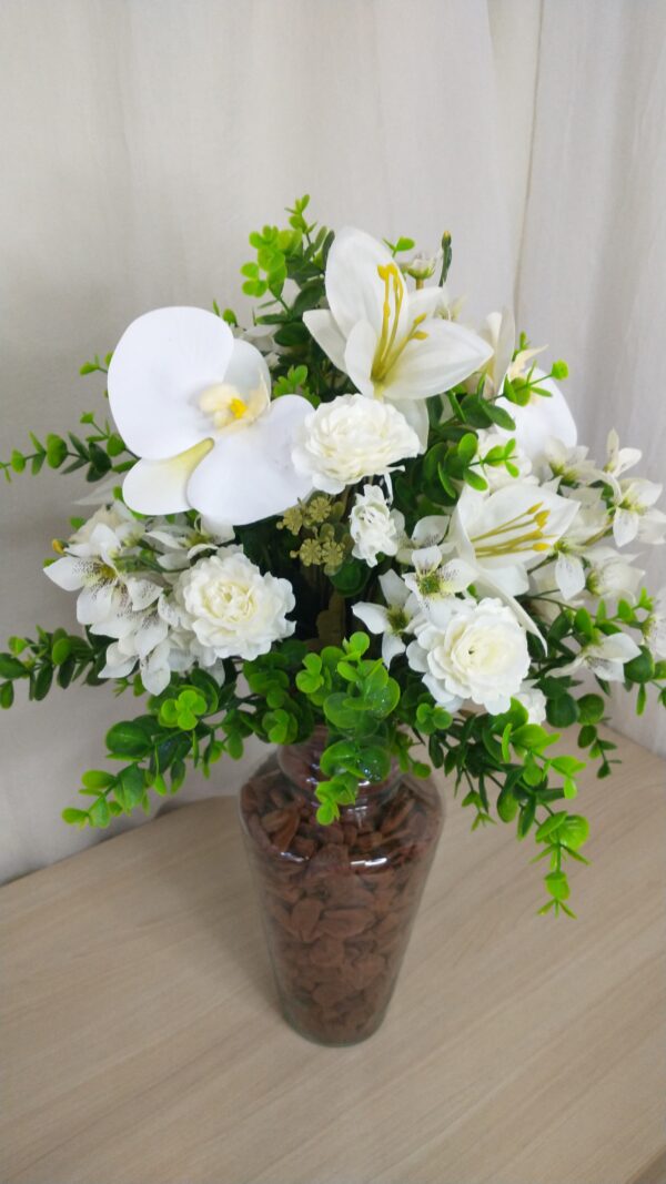 Vaso de vidro com arranjo de flores brancas, incluindo orquídeas, lírios e hortênsias, decorado com folhas verdes brilhantes.