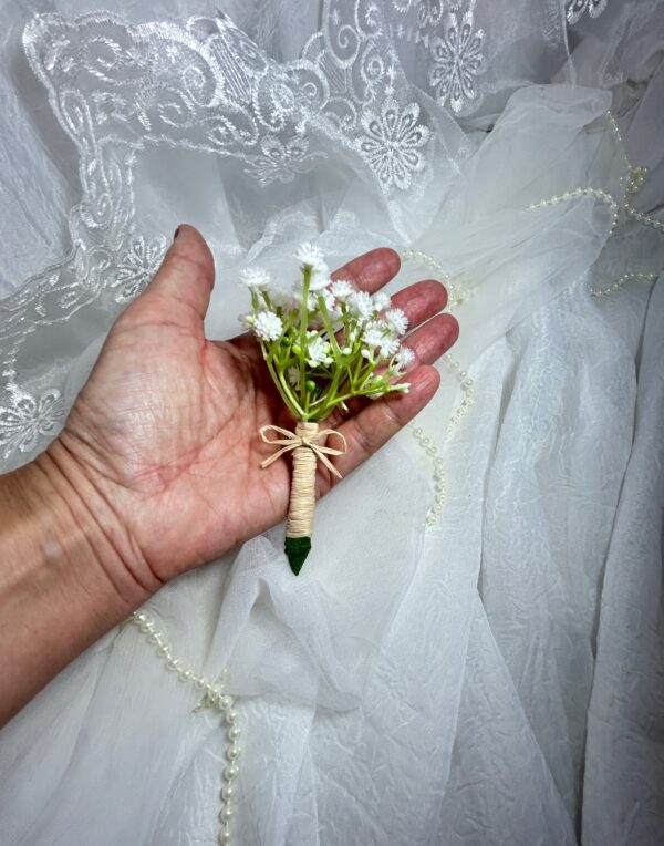Bouquê de flores com pequenos detalhes brancos e cabeça verde, mão que segura o arranjo, fundo de tecido branco e renda.